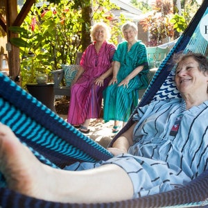 Op de afbeelding: Drie vrouwen ontspannen in een blauwe en turquoise gestreepte hangmat. Twee vrouwen zitten op een bank op de achtergrond en dragen roze en turquoise gebloemde jurken. De vrouw in de hangmat draagt een blauw-wit gestreepte jurk. De hangmat heeft een blauw-wit gebloemd kussen. De tekst "Bowerbird photography" is zichtbaar in de rechterbovenhoek van de afbeelding.