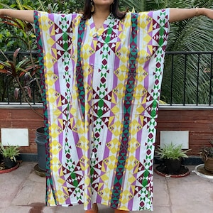 Puede incluir: Un vestido maxi blanco con un patrón geométrico en tonos de morado, amarillo, verde y burdeos. El vestido tiene cuello en V y mangas largas.