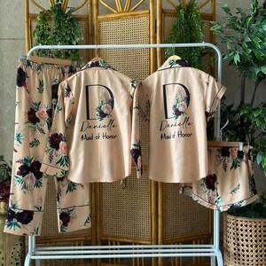 May include: Two matching sets of pajamas in a light beige color with a floral print. The pajamas include a long-sleeved shirt and pants. The shirts have a large letter "D" with a floral design and the text "Danielle Maid of Honor" printed on the front.