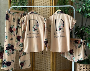May include: Two matching sets of pajamas in a light beige color with a floral print. The pajamas include a long-sleeved shirt and pants. The shirts have a large letter "D" with a floral design and the text "Danielle Maid of Honor" printed on the front.