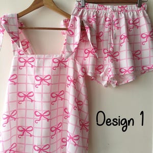 Puede incluir: Un conjunto de pijama de dos piezas con un estampado de lazo blanco y rosa. El conjunto incluye una parte superior con tirantes anudados y pantalones cortos con cintura con cordón. Las palabras "Design 1" están escritas en negro a la derecha.