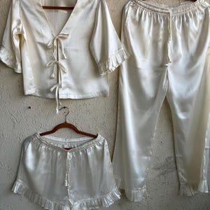 May include: An ivory satin pajama set comprising a short-sleeved top with tie fastenings, shorts, and cropped trousers. The set has frilled edges on the sleeves, shorts, and trousers. All items are displayed on wooden hangers against a neutral backdrop.