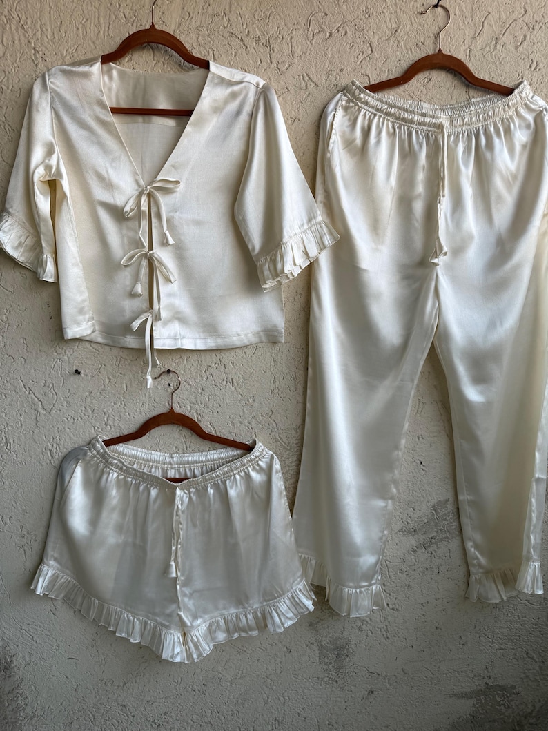 May include: An ivory satin pajama set comprising a short-sleeved top with tie fastenings, shorts, and cropped trousers. The set has frilled edges on the sleeves, shorts, and trousers. All items are displayed on wooden hangers against a neutral backdrop.