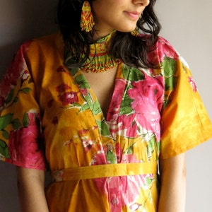 Può includere: Una donna indossa un abito giallo a fiori con una collana e orecchini di perline verdi e gialle. L'abito ha una scollatura a V e una chiusura con bottoni sul davanti.