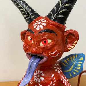 May include: A red devil figurine with white floral designs and a long blue tongue. The devil has large black horns with white accents, red eyes, and small blue and yellow wings. The figurine is a handcrafted piece.