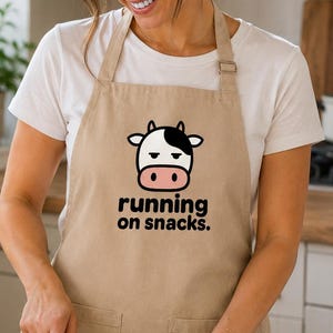 Puede incluir: Un delantal de color beige con un gráfico de una cara de vaca con expresión somnolienta y el texto "running on snacks". El delantal cuenta con una correa ajustable para el cuello con hebilla metálica y dos bolsillos frontales en la parte inferior.