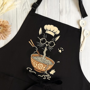Puede incluir: Delantal negro con un dise&ntilde;o de gato cocinero de dibujos animados. El gato lleva gafas y un gorro de chef, comiendo fideos ramen de un cuenco floral con palillos. El delantal tiene una correa ajustable para el cuello y lazos en la cintura.