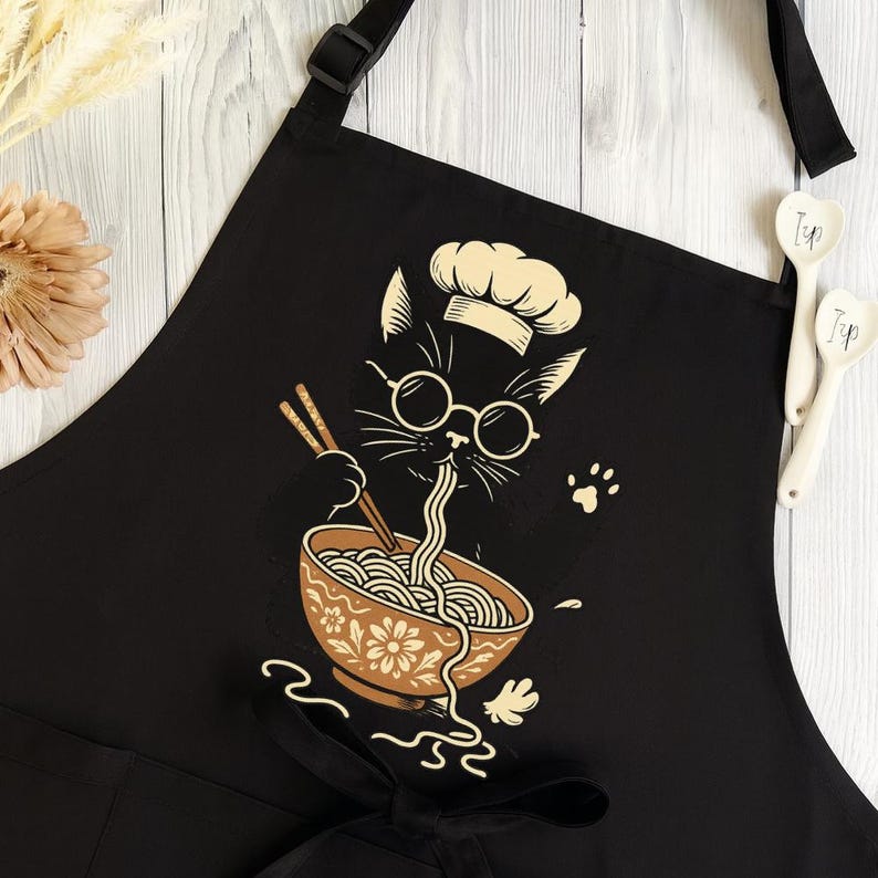 Puede incluir: Delantal negro con un dise&ntilde;o de gato cocinero de dibujos animados. El gato lleva gafas y un gorro de chef, comiendo fideos ramen de un cuenco floral con palillos. El delantal tiene una correa ajustable para el cuello y lazos en la cintura.