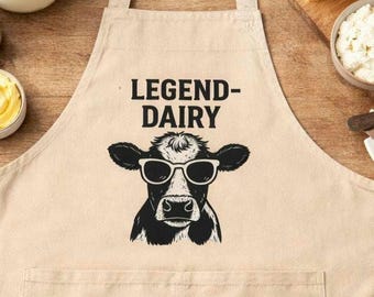 May include: Beige apron with the text "LEGEND-DAIRY" above a black and white illustration of a cow wearing sunglasses. The apron has a large front pocket and adjustable neck strap. The apron is on a wooden surface.