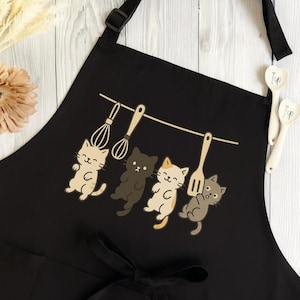 Pode incluir: Avental preto com um design lúdico de quatro gatos de desenho animado segurando utensílios de cozinha. Os gatos são em várias cores, incluindo bege, preto e cinza. Duas colheres de medição brancas estão no lado direito.