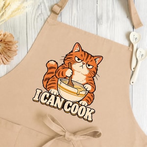 May include: A beige apron featuring a graphic of an orange tabby cat with a grumpy expression, stirring a bowl with a wooden spoon. Below the cat, the text I CAN COOK is printed in bold, blocky letters. The apron has adjustable neck straps.