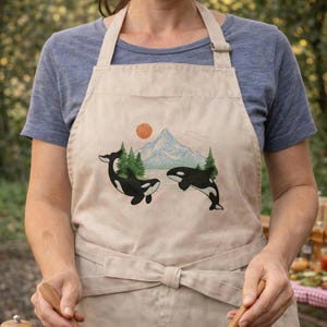 May include: Beige apron featuring a mountain scene with two black and white orcas, green trees, and a setting sun. The apron has a front pocket and a tie-waist design. The apron is made of a durable, stain-resistant fabric.