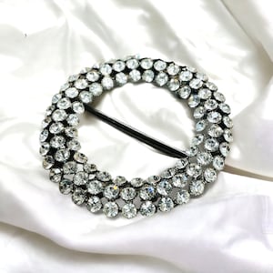 May include: A round, silver-toned buckle with a black bar. The buckle is encrusted with numerous clear, sparkling rhinestones, arranged in a circular pattern. The buckle is set against a white, silky background.
