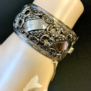 Puede incluir: Un brazalete de bisagra plateado con un diseño floral adornado. El brazalete tiene una cadena unida al cierre.