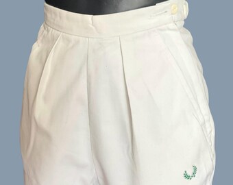 Könnte beinhalten: Weiße Shorts mit Falten und Knopfverschluss. Die Shorts haben ein kleines, grün gesticktes Emblem am Bein. Hergestellt aus leichtem Stoff.