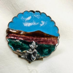 Könnte beinhalten: Vintage Brosche mit einer Landschaftsszene mit blauem Himmel, braunen Bergen und grünen und roten Feldern. Eine silberfarbene Elfenfigur sitzt im Vordergrund. Die Brosche hat einen gewellten Rand.