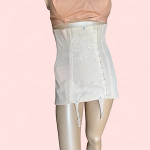 May include: Off-white vintage girdle with floral detailing and garter straps. The garment has a hook-and-eye closure and vertical boning for structure. The girdle is worn over a peach-coloured bra.