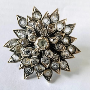 May include: A vintage-style brooch in the shape of a flower. The brooch features a central, round, clear stone surrounded by multiple clear stones. The petals are made of a gold-colored metal, and the brooch is set against a white background.