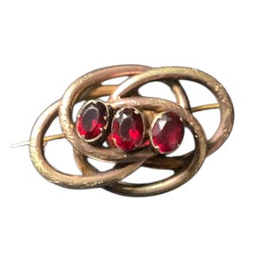 May include: Vintage gold-coloured brooch with three oval, deep red gemstones set in an ornate, intertwined design. The brooch has a pin closure, likely antique jewellery.