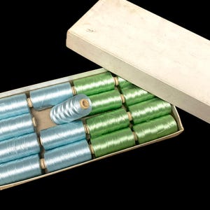 May include: A box of spools of thread in shades of blue and green. The spools are neatly arranged in a rectangular box with a lid. The thread appears to be a shiny, silky material, perfect for sewing or embroidery projects.