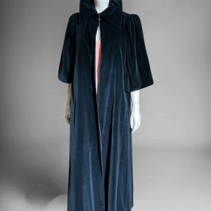 May include: A floor-length, dark teal velvet coat featuring a high, structured collar and wide, elbow-length sleeves. The coat is open at the front, revealing a glimpse of a light pink garment underneath, and has a smooth, matte texture.