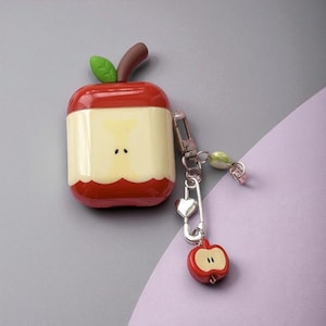 Könnte beinhalten: Ein rot-cremefarbenes, apfelförmiges Etui für kabellose Ohrhörer. Das Etui hat ein grünes Blatt und einen braunen Stiel. Ein silberner Schlüsselanhänger mit Herzanhänger und kleinem Apfelanhänger ist befestigt.