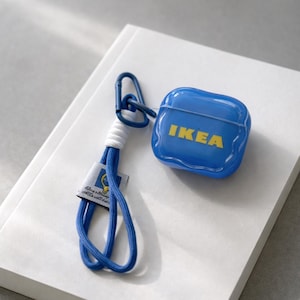 Puede incluir: Una funda azul para AirPods con el logotipo de IKEA en amarillo, sujeta a un cordón azul y blanco. El cordón tiene un mosquetón azul y una pequeña etiqueta con texto y un logotipo. La funda está sobre una superficie blanca.