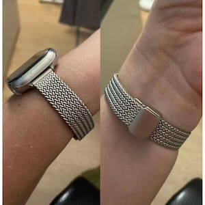 May include: A silver metal watch band with a woven mesh design. The band is shown on a person's wrist.