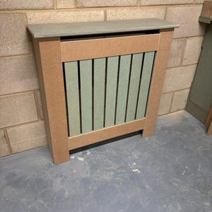 May include: A radiator cover with a light brown wood frame and a light green slatted front. The cover has a flat top.