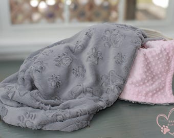May include: A gray and pink reversible pet blanket with a paw print design on one side and a textured dot pattern on the other side. The blanket is folded and displayed on a wooden surface. The brand name "Bark N Blankie" is visible in the lower right corner of the image.