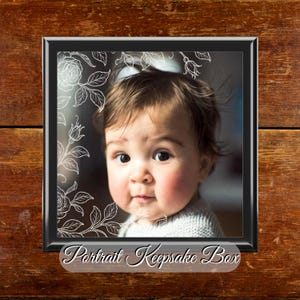 May include: A black framed portrait keepsake box featuring a baby's photograph. The frame is decorated with a floral pattern. The text "Portrait Keepsake Box" is displayed at the bottom. The background is a wooden surface.