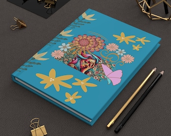 May include: A teal journal with a colorful floral and butterfly design. The cover features a detailed illustration of a woman's face surrounded by flowers and a pink butterfly. Gold and black pencils and gold binder clips are also present.