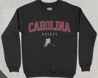Pode incluir: Camisola de moletom preta com um logotipo Carolina Hockey vermelho e branco. O logotipo apresenta um jogador de hóquei patinando com um taco. O texto "CAROLINA" está em uma fonte grande e em negrito acima do texto "HOCKEY".