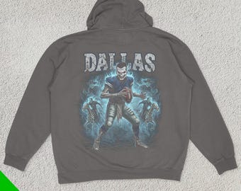 May include: A dark grey hooded sweatshirt with a graphic print of a football player in a blue and white uniform. The player is holding a football and is surrounded by a blue and white lightning bolt design. The text "DALLAS" is printed in white above the player.