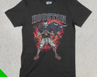 Op de afbeelding: Zwart T-shirt met een afbeelding van een skelet-voetballer die een voetbal vasthoudt en een stier met vlammen erachter. De tekst "HOUSTON" staat boven de afbeelding.