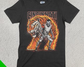 Può includere: Maglietta nera con una grafica di una tigre e un giocatore di football in fiamme. Il testo "Cincinnati" è sopra la grafica.