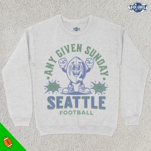 May include: A grey sweatshirt with a cartoon football character and the text "Any Given Sunday. Seattle Football. Est'd 1976."