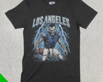 Op de afbeelding: Zwart t-shirt met een skeletachtige footballspeler in een blauw en wit uniform, met een football. De woorden "LOS ANGELES" staan boven de speler, met bliksemflitsen op de achtergrond. Het shirt heeft een Fan Ghoul logo.