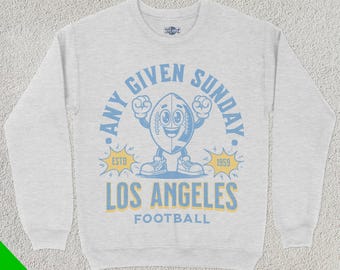 May include: A grey sweatshirt with a blue and yellow graphic design. The graphic features a cartoon football with a smiling face and the text "Any Given Sunday" and "Los Angeles Football".