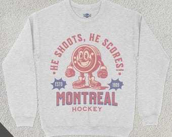Puede incluir: Una sudadera gris con un gráfico rojo y blanco de un disco de hockey con una cara. El gráfico dice "¡Él dispara, él anota!" con el texto "Montreal Hockey" debajo. El texto "Estd 1909" está en forma de estrella.
