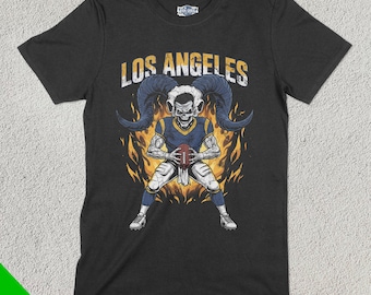 Könnte beinhalten: Schwarzes T-Shirt mit einer Grafik eines skelettartigen Footballspielers mit großen Hörnern, der einen Football hält. Der Text "LOS ANGELES" befindet sich über der Figur, mit Flammen im Hintergrund. Das Shirt hat das "Fan Ghoul"-Logo.