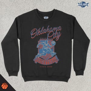 Może przedstawiać: Czarna bluza z nadrukiem w stylu vintage przedstawiającym koszykarza przed tarczą z napisem "Oklahoma City Thunder" i "Since 2008".