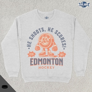 May include: A grey sweatshirt with a vintage-style hockey logo. The logo features a cartoon puck with a smiling face and the text "He shoots, he scores!"  The text "Edmonton Hockey" is below the logo.