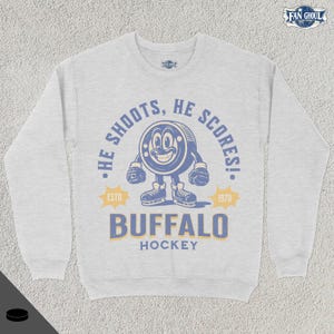 May include: A gray sweatshirt with a vintage-style hockey graphic. The graphic features a cartoon puck with a face and the text "He shoots, he scores!" with "ESTD 1970" below. The text "BUFFALO HOCKEY" is printed in a large font below the graphic.