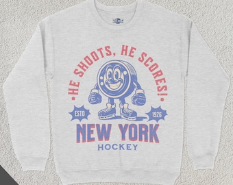 May include: A gray sweatshirt with a vintage-style graphic of a hockey puck with a face. The graphic says "He Shoots, He Scores!" with "ESTD 1926" and "NEW YORK HOCKEY" below it.