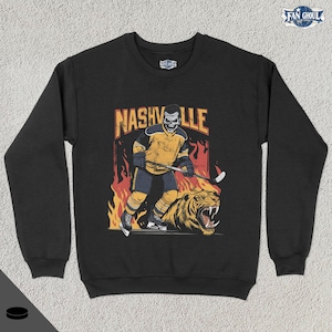 May include: Black sweatshirt with a graphic of a hockey player wearing a yellow and black jersey with the word "NASHVILLE" above him. The player is holding a hockey stick and is standing on a tiger.