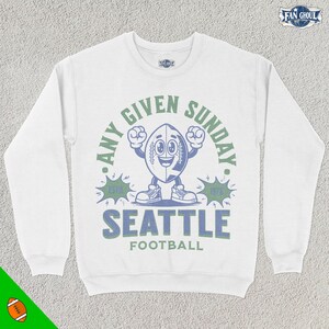 May include: White long-sleeve sweatshirt with a blue and green graphic of a football character with the text "Any Given Sunday. Seattle Football. Est. 1976."