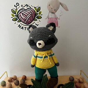 May include: A crocheted raccoon wearing a yellow and green sweater and green pants. The raccoon is standing on a wooden shelf with a green leafy vine and brown and gold striped balls.