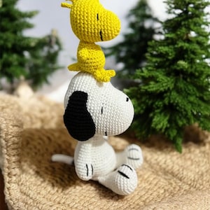 Pode incluir: Figuras de croch&ecirc; de Snoopy e Woodstock. Snoopy &eacute; branco com detalhes pretos, e Woodstock &eacute; amarelo. Est&atilde;o sobre uma superf&iacute;cie bege, com &aacute;rvores verdes ao fundo. As figuras s&atilde;o feitas &agrave; m&atilde;o e parecem macias.