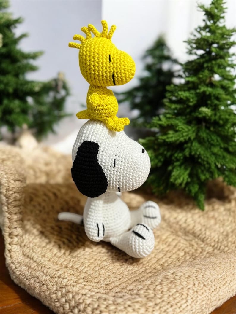 Pode incluir: Figuras de croch&ecirc; de Snoopy e Woodstock. Snoopy &eacute; branco com detalhes pretos, e Woodstock &eacute; amarelo. Est&atilde;o sobre uma superf&iacute;cie bege, com &aacute;rvores verdes ao fundo. As figuras s&atilde;o feitas &agrave; m&atilde;o e parecem macias.
